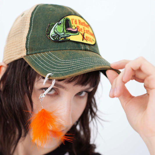 I'D RATHER BE EATING BASS FISH HOOK CAP, close-up on a model holding the brim; silver-tone hook and orange feather hang down