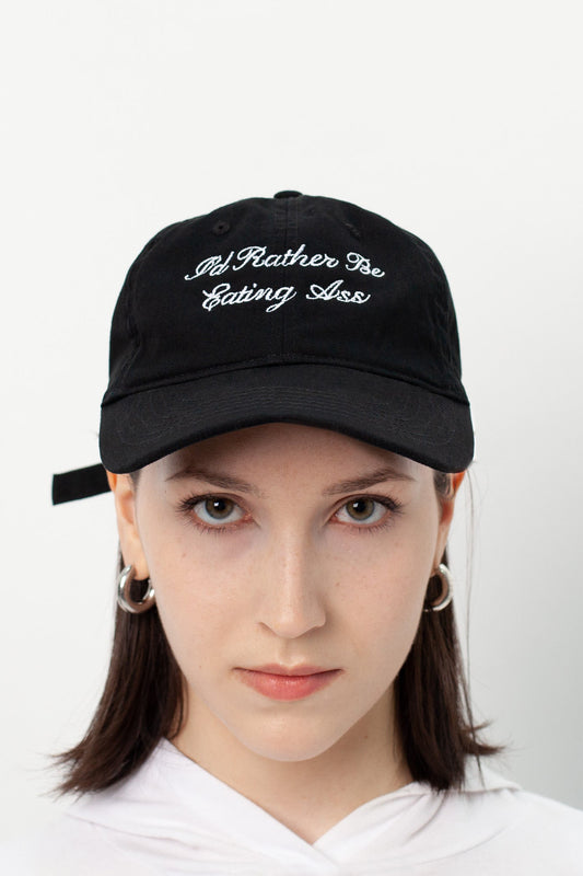 I'D RATHER BE EATING ASS CURSIVE BASEBALL CAP front view on a model: black cap with white cursive embroidery on the crown
