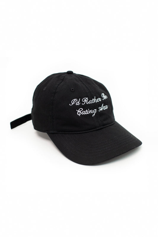 I'D RATHER BE EATING ASS CURSIVE BASEBALL CAP angled side view on white: black cap with white cursive embroidery, back strap