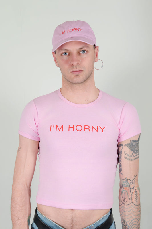 I'M HORNY T-SHIRT on a model, front view: light pink baby tee with red “I'M HORNY” graphic, worn with the matching cap