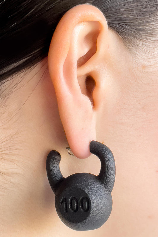 KETTLEBELL EARRINGS, tighter crop on the ear: black kettlebell marked "100" with its handle wrapped around the lobe